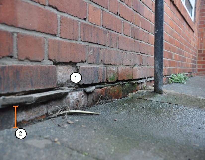 A photograph of a red brick wall on a rainy day. Wet concrete paving slabs are in contact with the wall, only one brick below the slate damp-proof course.