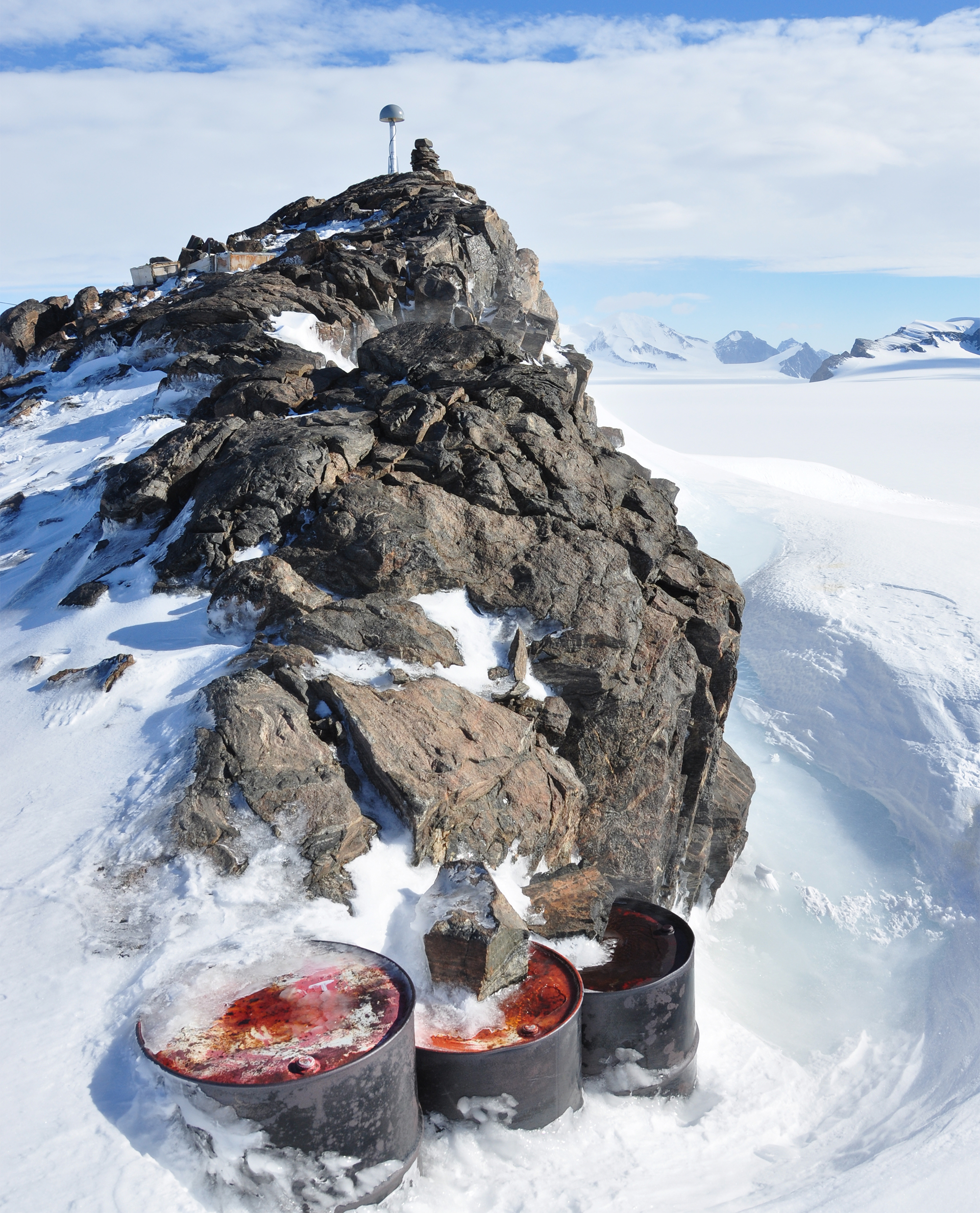 GNSS antenna and monument with aircraft fuel drums in the foreground at Jensen Nunatak in the Antarctic Peninsula