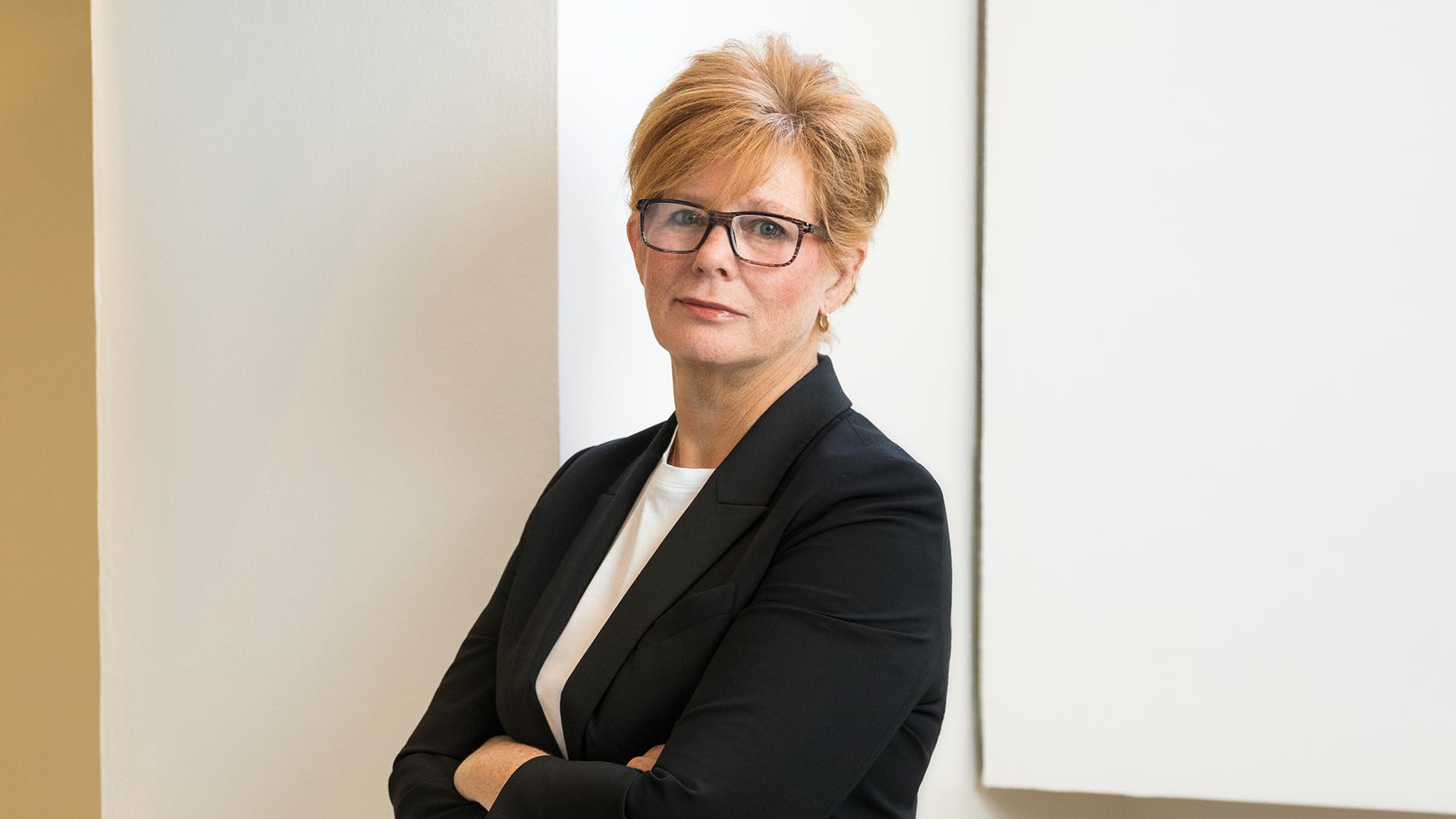 Ann Gray leaning against a wall with arms folded looking at camera