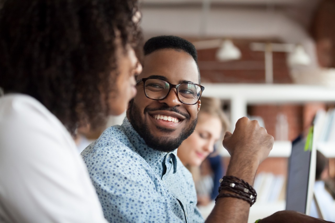 Close up of smiling African American employee look at female colleague chatting in office, happy black male worker talk with woman coworker, having casual conversation at workplace, have fun; Shutterstock ID 1209553408; Purchase Order: n/a; Client/Licensee: RICS