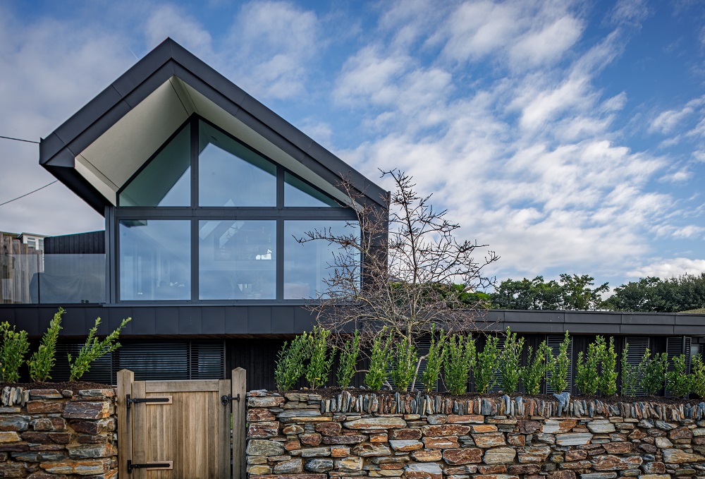 Image of house with sloped roof and glass windows, with stonewall fence in front and blue sky above